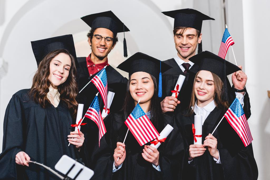 アメリカの旗を持ち、スマートフォンで自分撮りをする卒業ガウンで幸せな学生の選択的な焦点  - 写真・画像