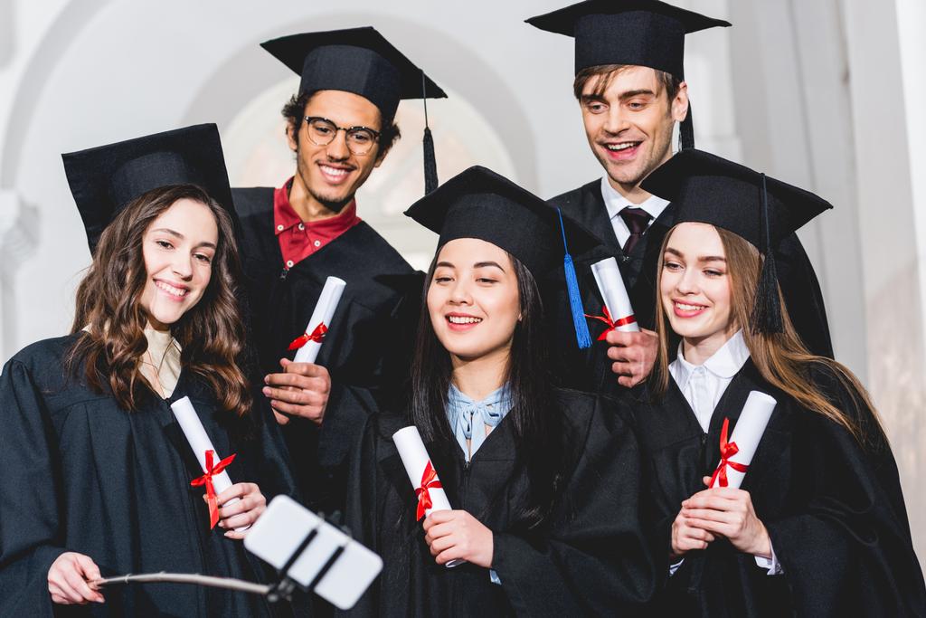 Wesoła grupa studentów w suknie dyplomowe posiadania dyplomów podczas rozmowy selfie  - Zdjęcie, obraz