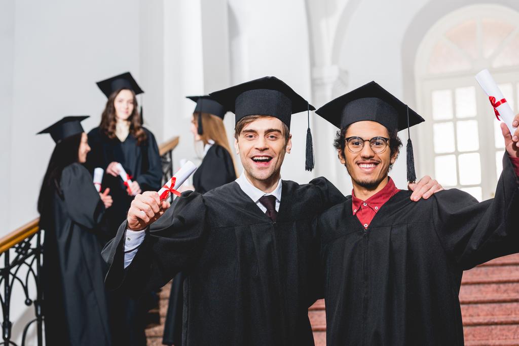 学生の近くに卒業証書を保持する卒業キャップで陽気な男性の選択的な焦点  - 写真・画像