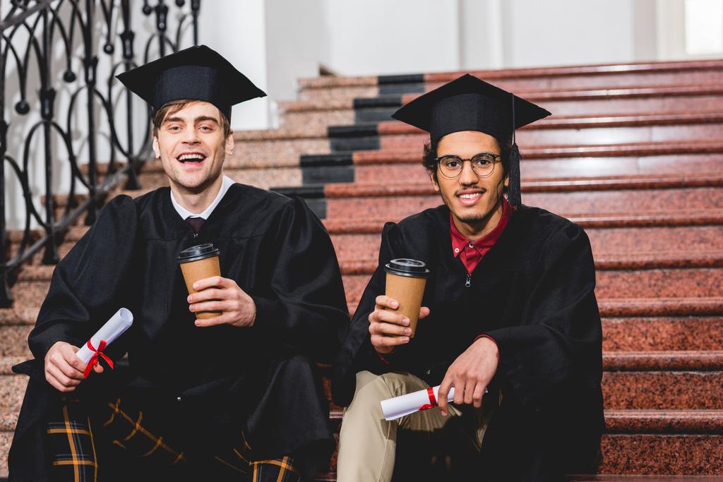 веселі студенти у випускних шапках, що тримають дипломи та паперові чашки
  - Фото, зображення
