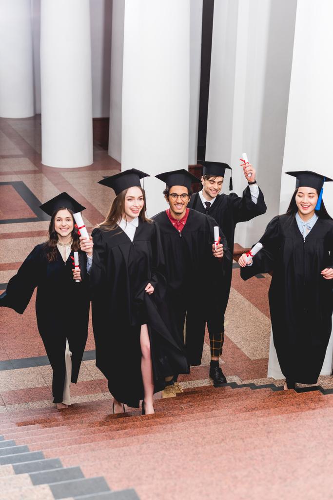estudantes alegres em vestidos de graduação segurando diplomas e andando em escadas
  - Foto, Imagem