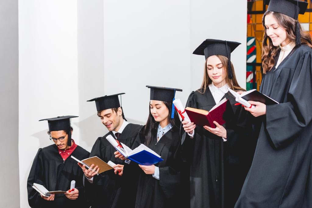 cheerful students in graduation gowns holding diplomas and reading books - Photo, Image