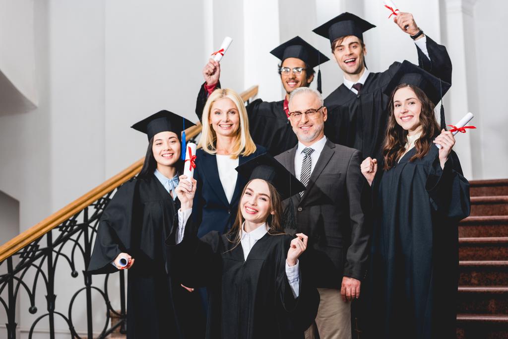 estudiantes alegres en gorras de graduación con diplomas cerca de profesores felices
  - Foto, imagen