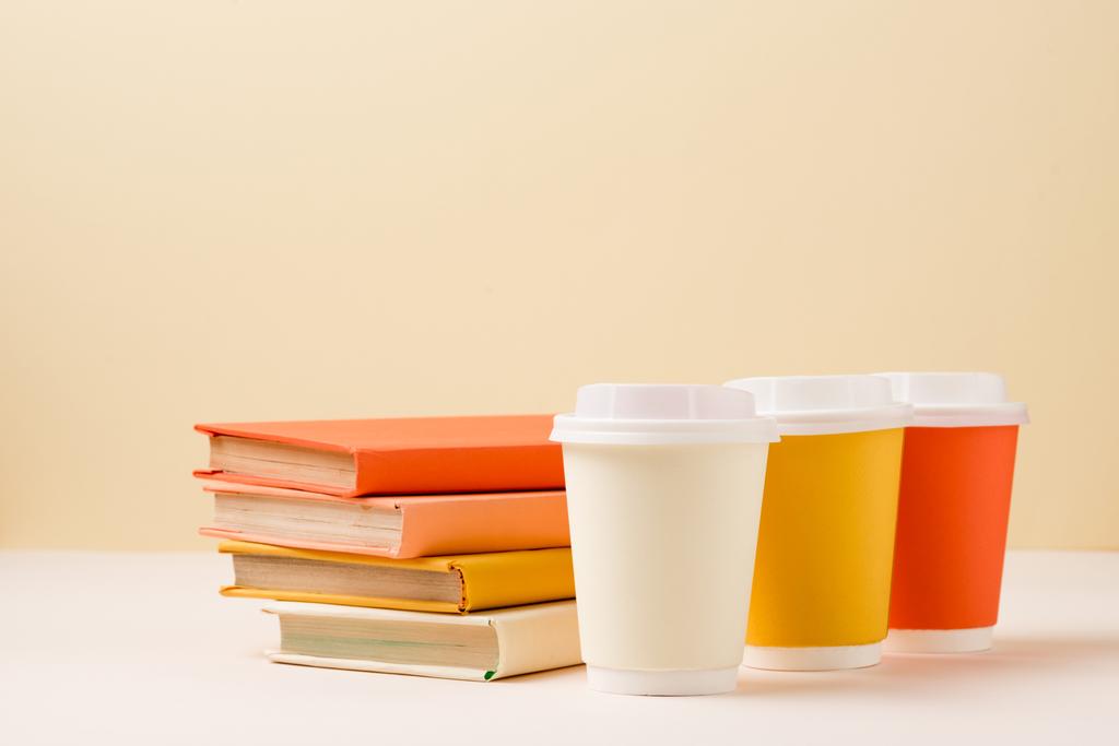 στοίβα βιβλίων και πολύχρωμα κύπελλα μίας χρήσης σε λευκή επιφάνεια απομονωμένη σε μπεζ - Φωτογραφία, εικόνα