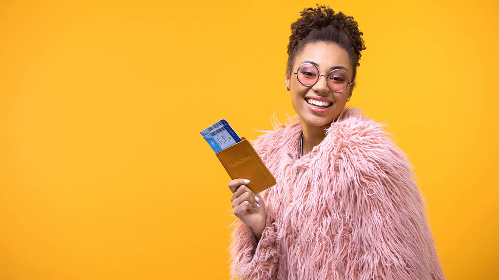 黄色の背景にパスポートと旅行チケットを示す笑顔のアフリカの女性 - 写真・画像