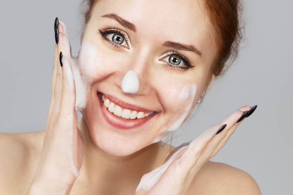 Portrait of cheerful laughing girl applying foam for washing on her face. Lovely woman redhead with attractive appearance. Skin Care spa relax concept on grey background - Photo, Image
