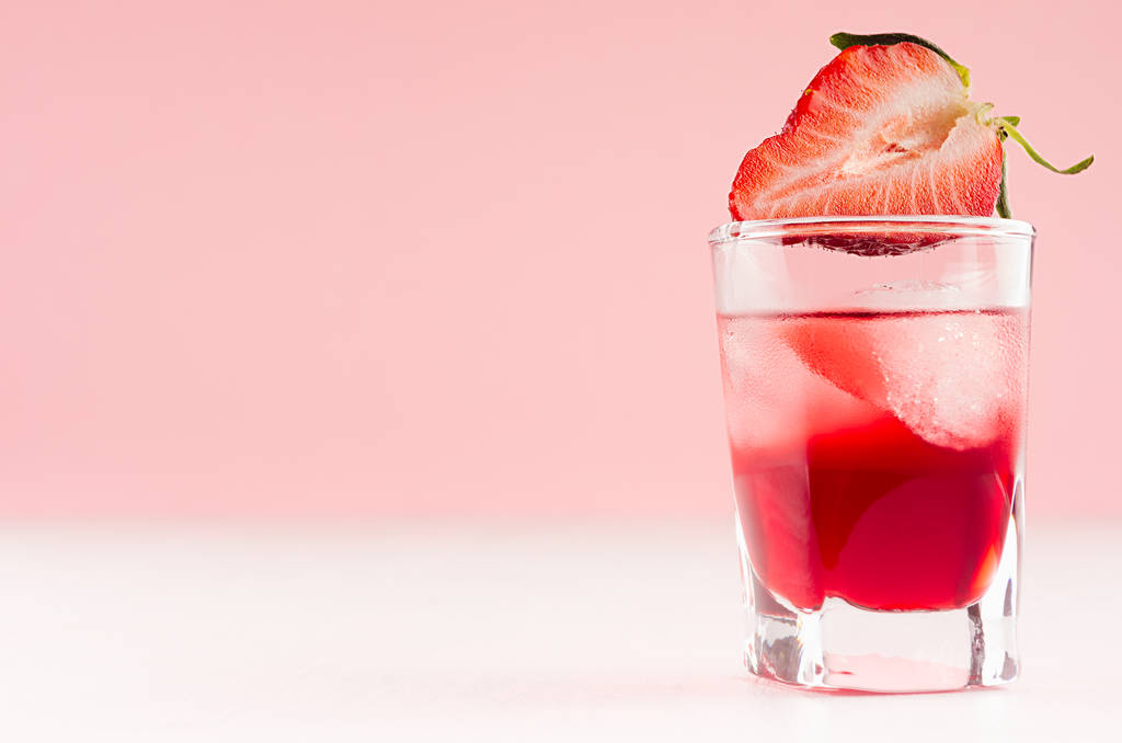 exquisite kalte rote alkoholische Spirituose mit Eiswürfeln und Erdbeerscheibe in Schnapsglasnahaufnahme auf weißem Holztisch und pastellrosa Wand, Kopierraum. - Foto, Bild