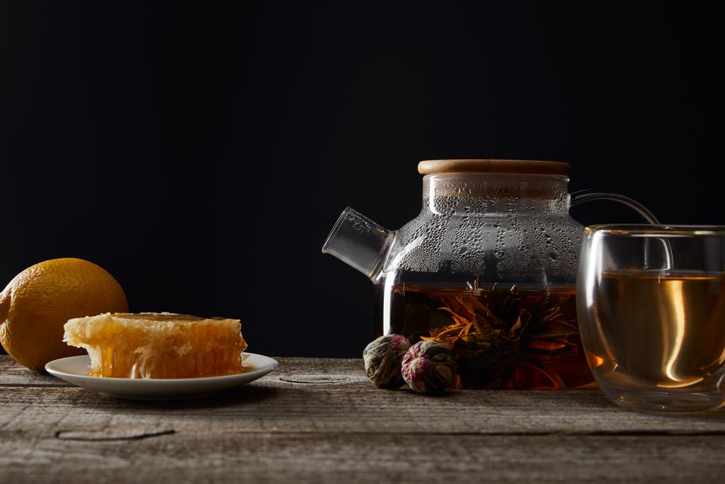 transparent teapot with blooming tea, glass, lemon and honeycomb on wooden table isolated on black - Photo, Image