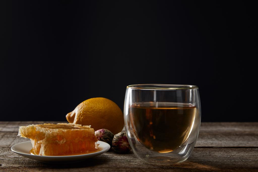 transparent glass with blooming tea, honeycomb and lemon on wooden table isolated on black - Photo, Image