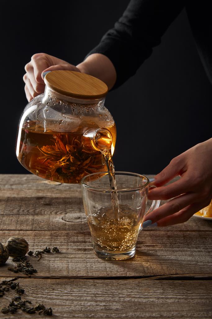 abgeschnittene Ansicht der jungen Frau gießt blühenden Tee aus Teekanne in Tasse isoliert auf schwarz - Foto, Bild
