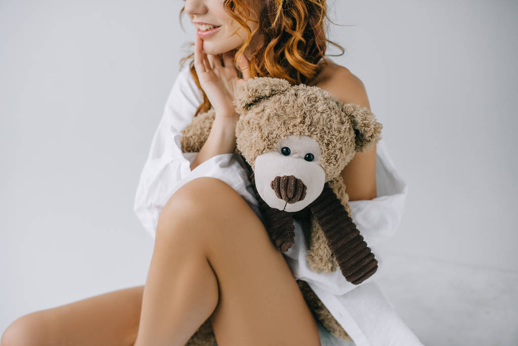 abgeschnittene Ansicht von fröhlich lockige junge Frau hält Teddybär, während sie auf weiß sitzt  - Foto, Bild