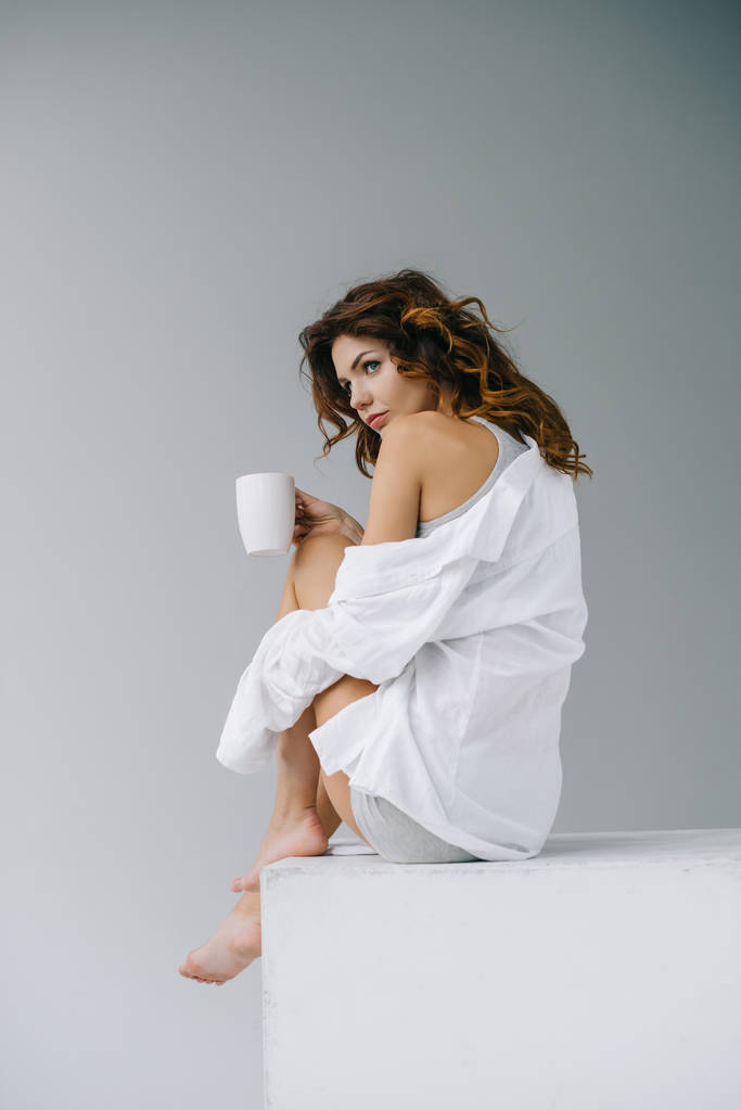 コーヒーとコーヒーと灰色の上に座ってカップを保持している魅力的な若い女性  - 写真・画像