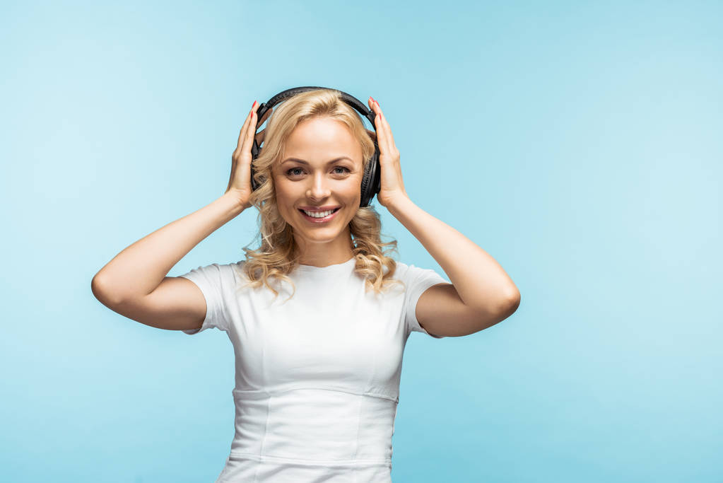 χαρούμενη ξανθιά γυναίκα ακούγοντας μουσική και αγγίζοντας τα μαύρα ακουστικά στο μπλε  - Φωτογραφία, εικόνα