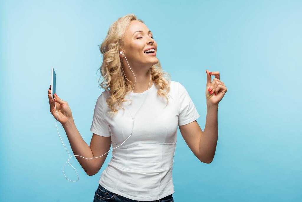 happy woman with closed eyes listening music in earphones while holding smartphone on blue  - Photo, Image
