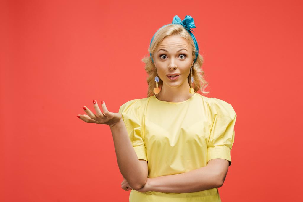 verrast blonde vrouw in blauwe hoofdband kijken naar camera en gebaren op rood  - Foto, afbeelding