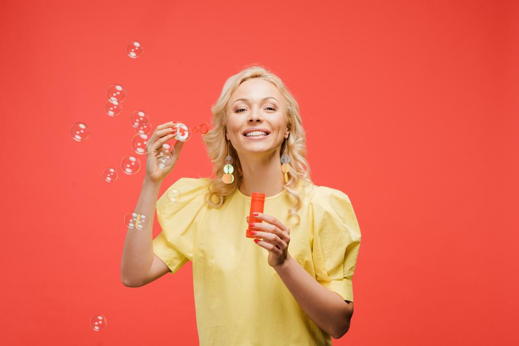cheerful blonde woman holding bottle near soap bubbles and smiling on red  - Photo, Image