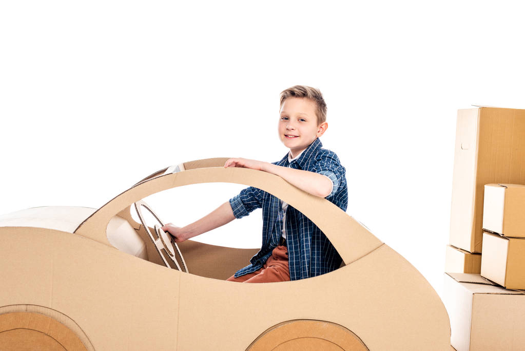 Улыбающийся мальчик, сидящий в картонной машине и смотрящий на камеру изолированный на белом
 - Фото, изображение