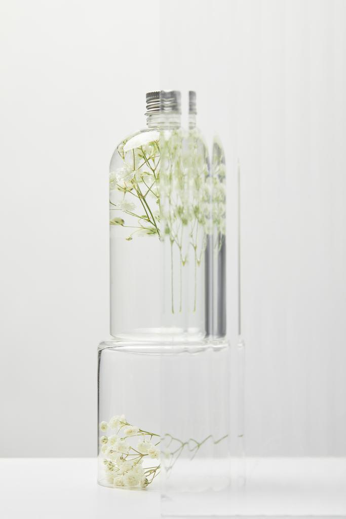 οργανικό καλλυντικό προϊόν σε διάφανη φιάλη με αγριολούλουδα πίσω από γυαλί σε λευκό τραπέζι σε γκρίζο φόντο - Φωτογραφία, εικόνα
