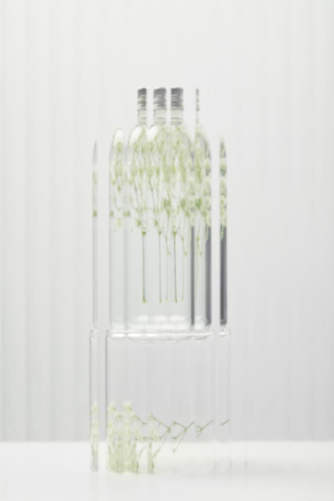 verschwommenes Bio-Kosmetikprodukt in transparenter Flasche mit Wildblumen hinter Glas auf grauem Hintergrund - Foto, Bild