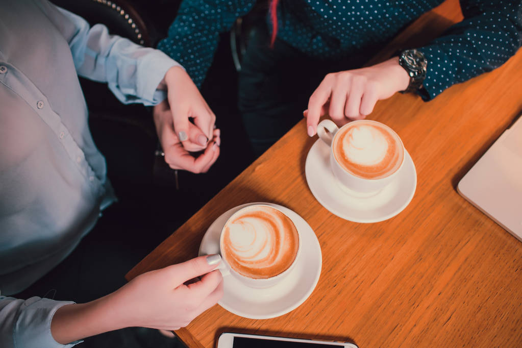 カフェの木製テーブルにコーヒーカップを持つ2人の手のクローズアップ画像. - 写真・画像