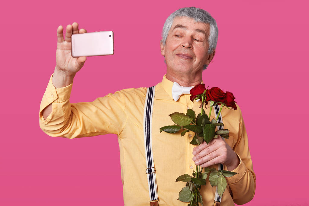 Cose up portrait of handsome senior man making selfie before dating, looks smiling at camera, wears yellow shirt with suspenders and bowtie, holding red roses, isolated over pink background. - Photo, Image