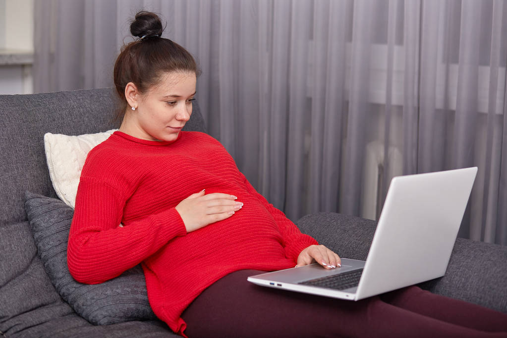Закритий постріл вагітної жінки одягнений червоний светр і бордовий легінси, тримає руку на животі, Годинники фільми онлайн, використовує бездротовий Інтернет, моделі в сучасній квартирі, сидячи босоніж критий. - Фото, зображення