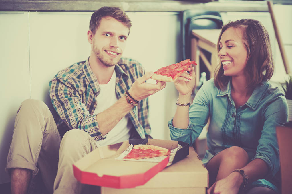 Giovani coppie hanno una pausa pranzo pizza sul pavimento dopo essersi trasferiti in una nuova casa con scatole intorno a loro
 - Foto, immagini