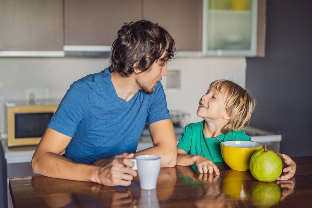 Πατέρας και γιος μιλάμε και χαμογελώντας ενώ έχοντας ένα πρωινό στην κουζίνα - Φωτογραφία, εικόνα