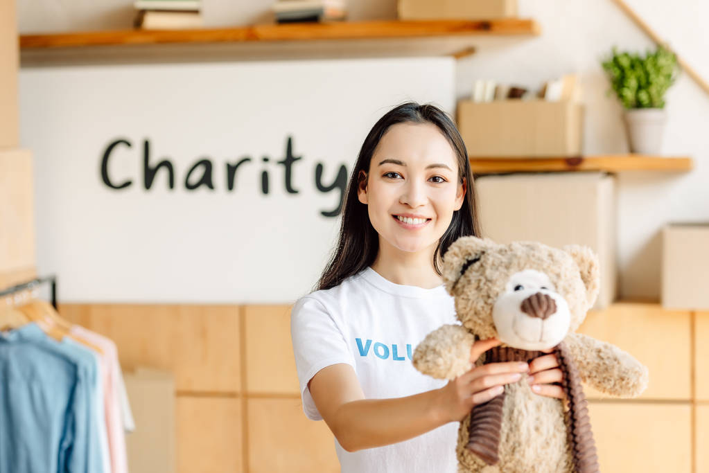 sorridente asiatico volontario ragazza mostrando orsacchiotto a macchina fotografica mentre in piedi vicino cartello con iscrizione di carità
 - Foto, immagini