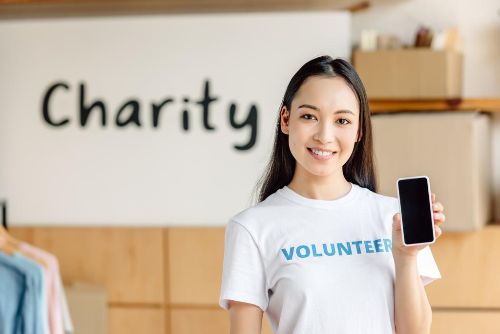 schöne asiatische Freiwillige hält Smartphone mit leerem Bildschirm, lächelt und schaut in die Kamera - Foto, Bild