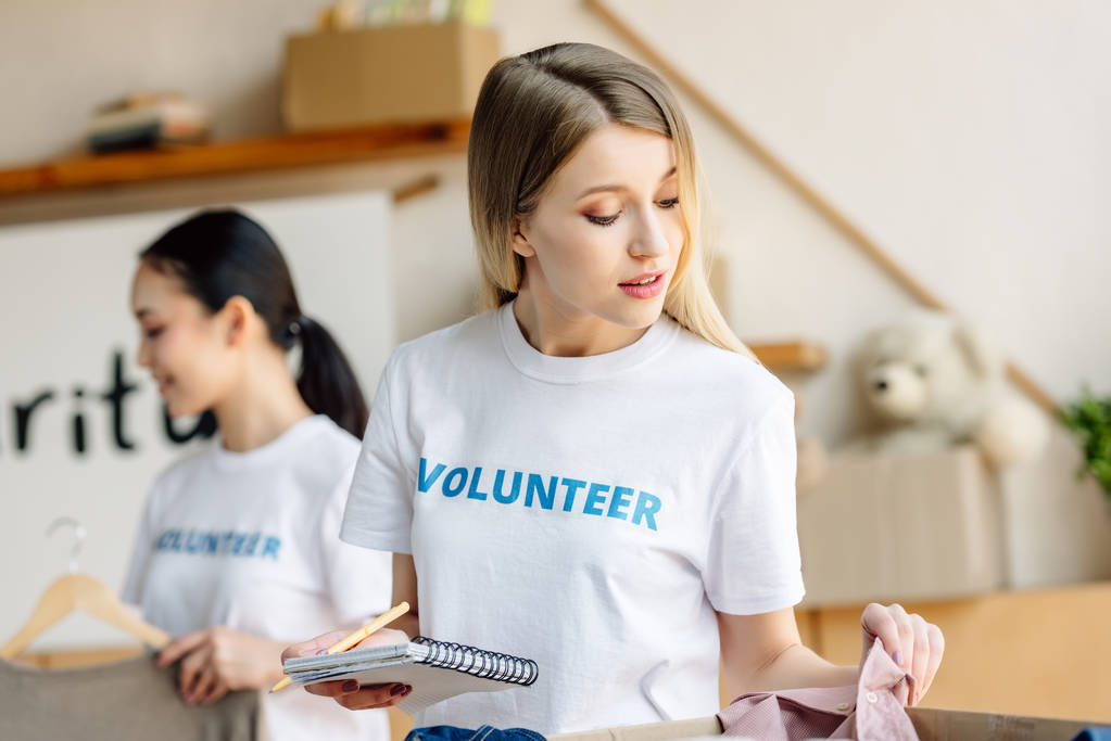 επιλεκτική εστίαση του ελκυστικού νεαρού εθελοντή γράφοντας σε φορητό υπολογιστή κοντά ασιάτισσα κορίτσι στέκεται με ράφι με ρούχα - Φωτογραφία, εικόνα