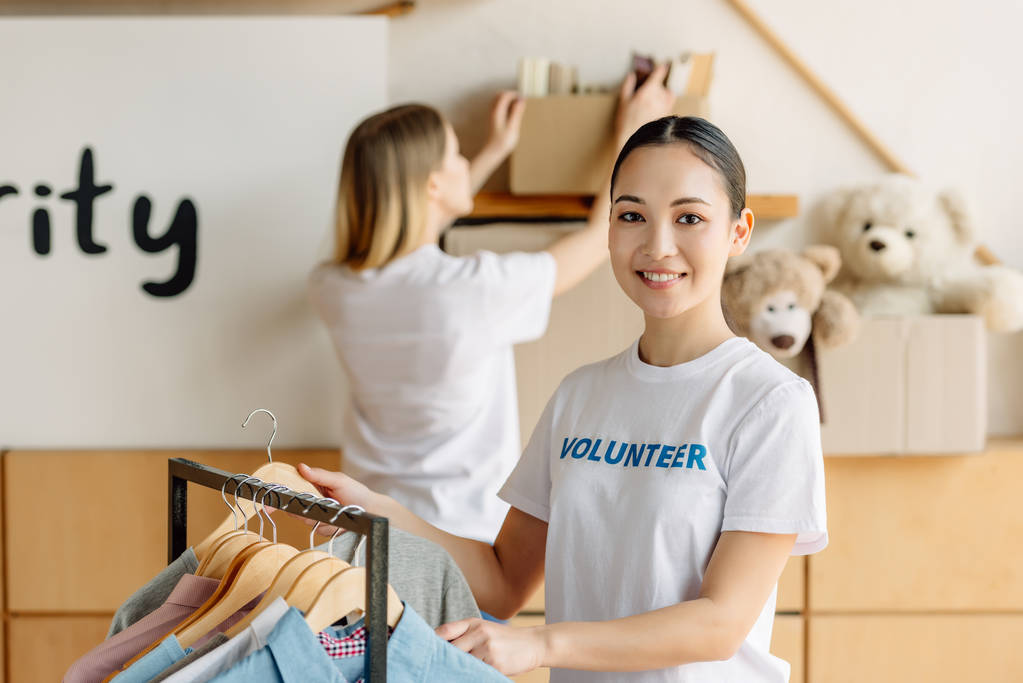 selective focus of pretty asian volunteer standing near rack with shirts and looking at camera - Photo, Image