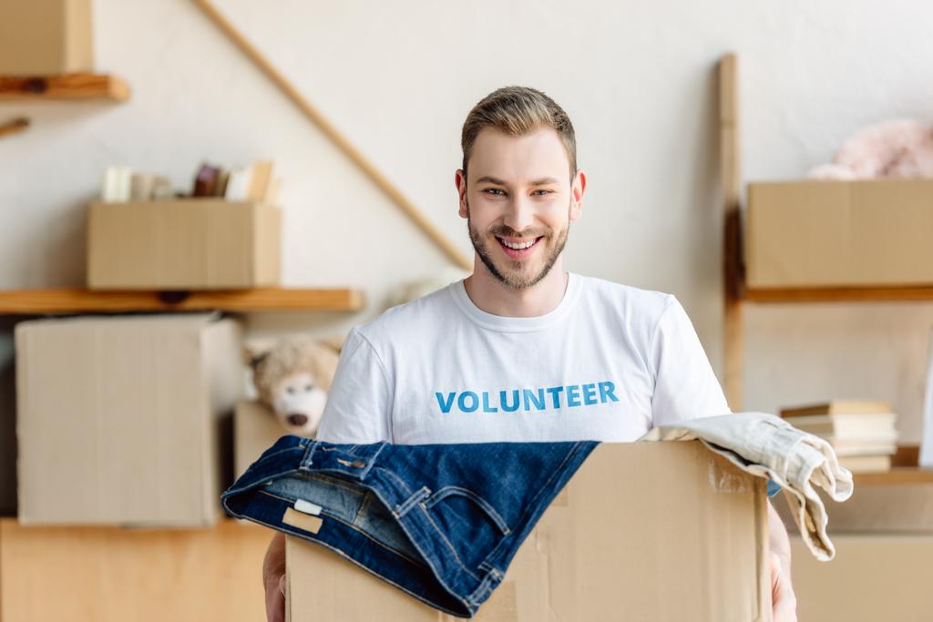 handsome young volunteer holding carton box with clothes, smiling and looking at camera - Photo, Image