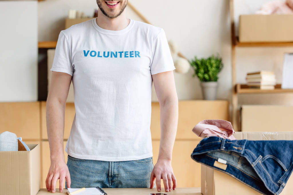 обрезанный вид добровольца, стоящего рядом с картонными коробками с одеждой
 - Фото, изображение
