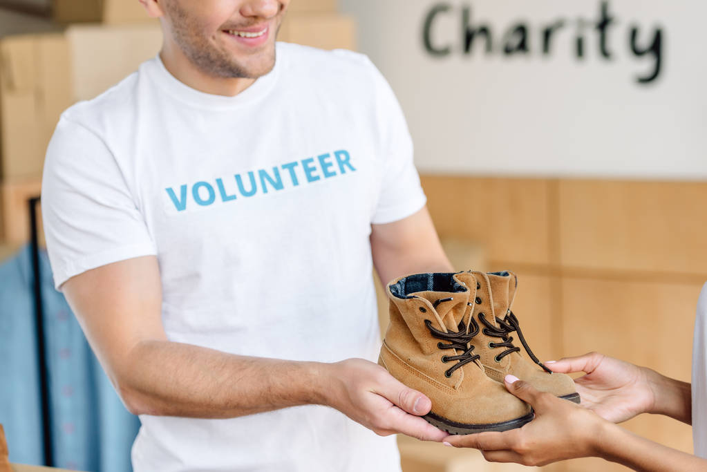 cropped view of two multicultural volunteers holding childrens shoes  - Photo, Image