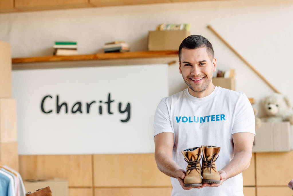 guapo, alegre voluntario sosteniendo los zapatos de los niños y mirando a la cámara
 - Foto, Imagen