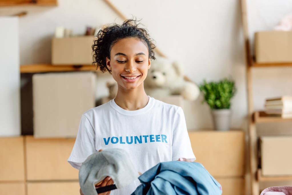 hermosa chica afroamericana en camiseta blanca con inscripción voluntaria sosteniendo ropa
  - Foto, Imagen