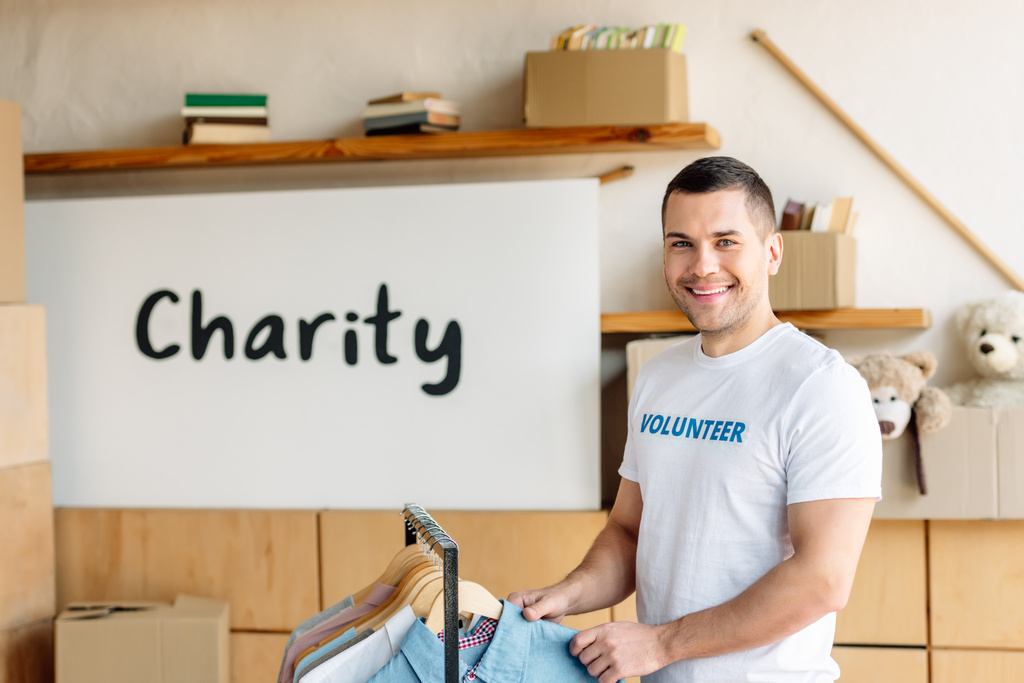 όμορφος, χαρούμενος εθελοντής στέκεται κοντά σε rack με πουκάμισα και κοιτάζοντας την κάμερα - Φωτογραφία, εικόνα