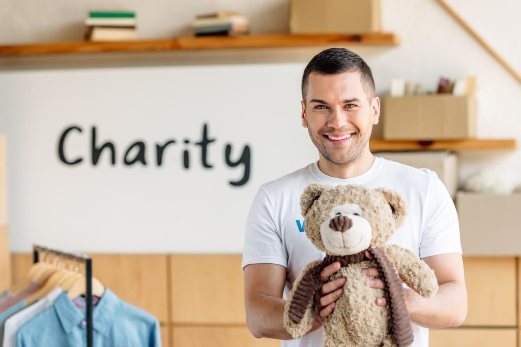 handsome, cheerful volunteer holding teddy bear and looking at camera  - Photo, Image