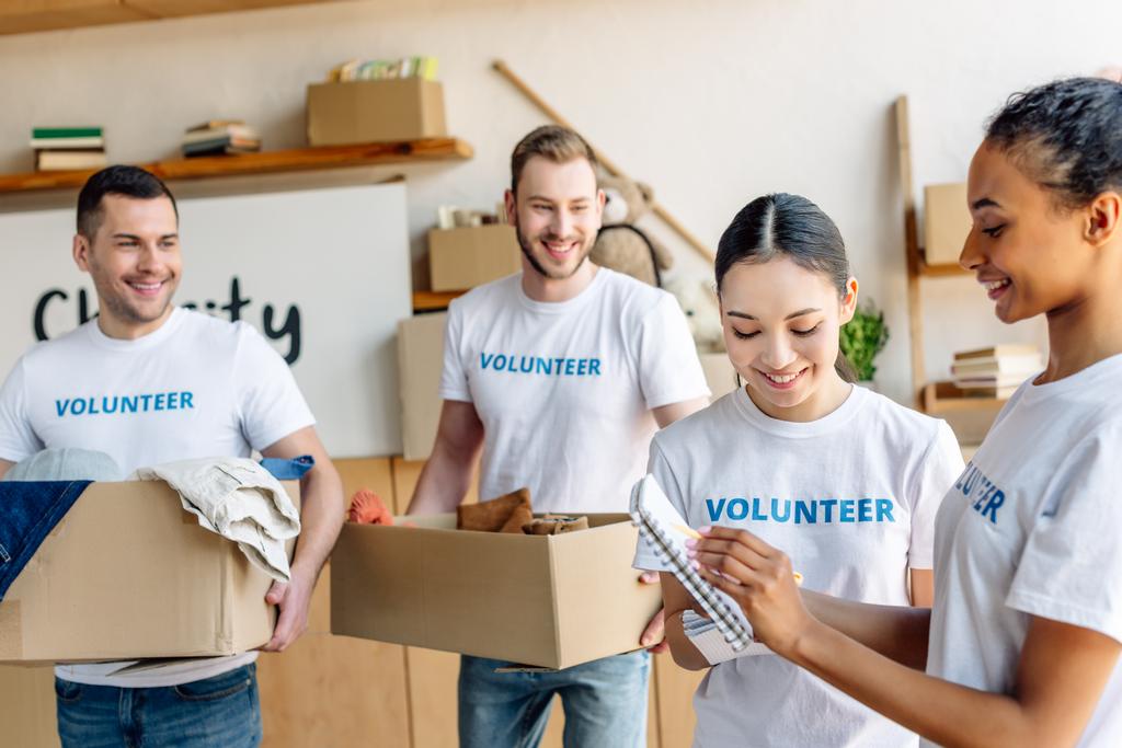 четыре молодых мультикультурных волонтера в белых футболках с волонтерскими надписями, работающих в благотворительном центре
 - Фото, изображение