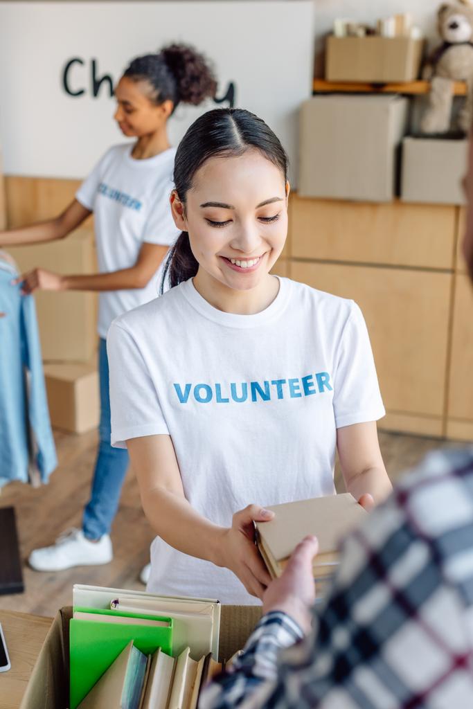 selektivní zaměření usmívajícího se asijského dobrovolníka, který dával knihy člověku zatímco africká americká dívka stojící blízko stojanu s košilí - Fotografie, Obrázek