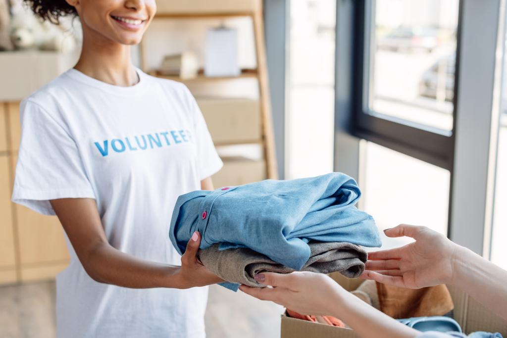 vista cortada de Africano americano voluntário dando roupas para a mulher no centro de caridade
 - Foto, Imagem