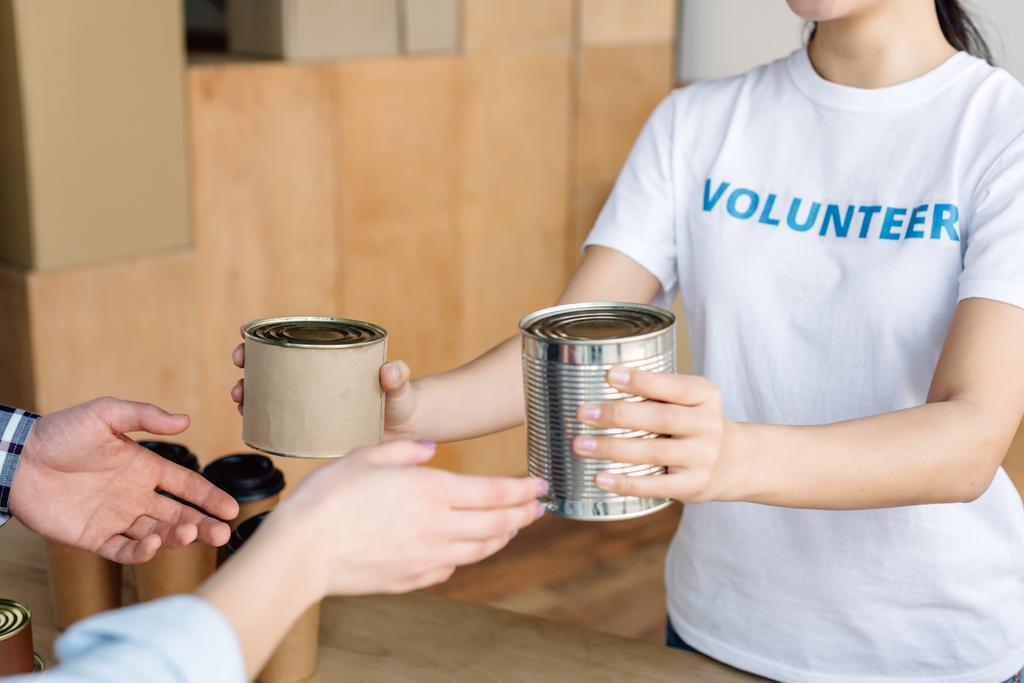 bijgesneden overzicht van vrijwilligers geven van ingeblikt voedsel aan man en vrouw in liefdadigheids centrum - Foto, afbeelding