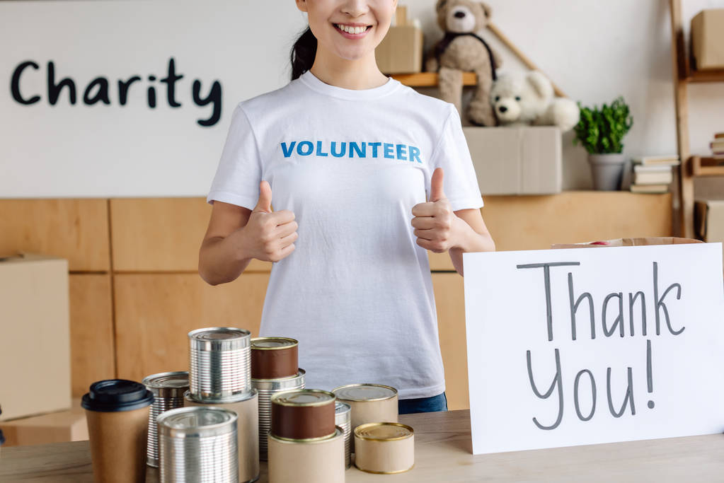 przycięte widok wolontariuszy pokazując kciuki w górę stojąc przy stole z konserwy i karty z Dziękuję napis - Zdjęcie, obraz