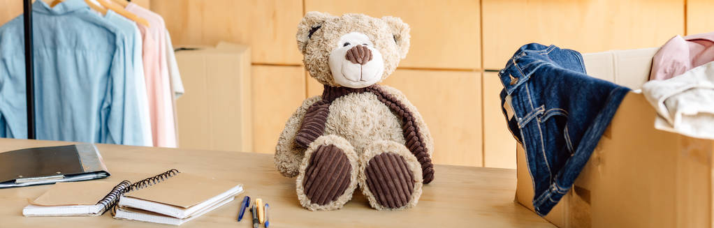 Panoramaaufnahme von Holztisch mit Teddybär, Karton mit Kleidung und Notizbüchern - Foto, Bild