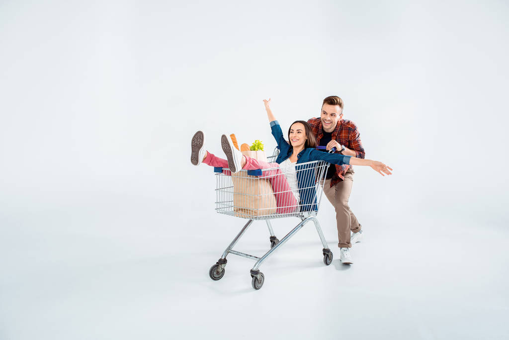 Šťastný mladý muž tlačí nákupního košíku s vzrušená žena a s potravinami pytel na bílém - Fotografie, Obrázek
