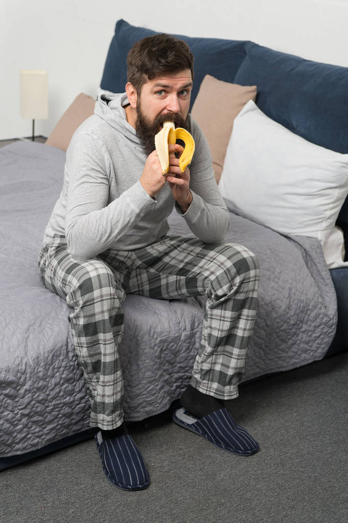 Vibrazioni mattutine a casa. hipster uomo barbuto mangiare banana al mattino. brutale uomo assonnato fa colazione in camera da letto. maschio maturo con barba in pigiama sul letto. dormire e svegliarsi. stile di vita sano. dolce casa - Foto, immagini
