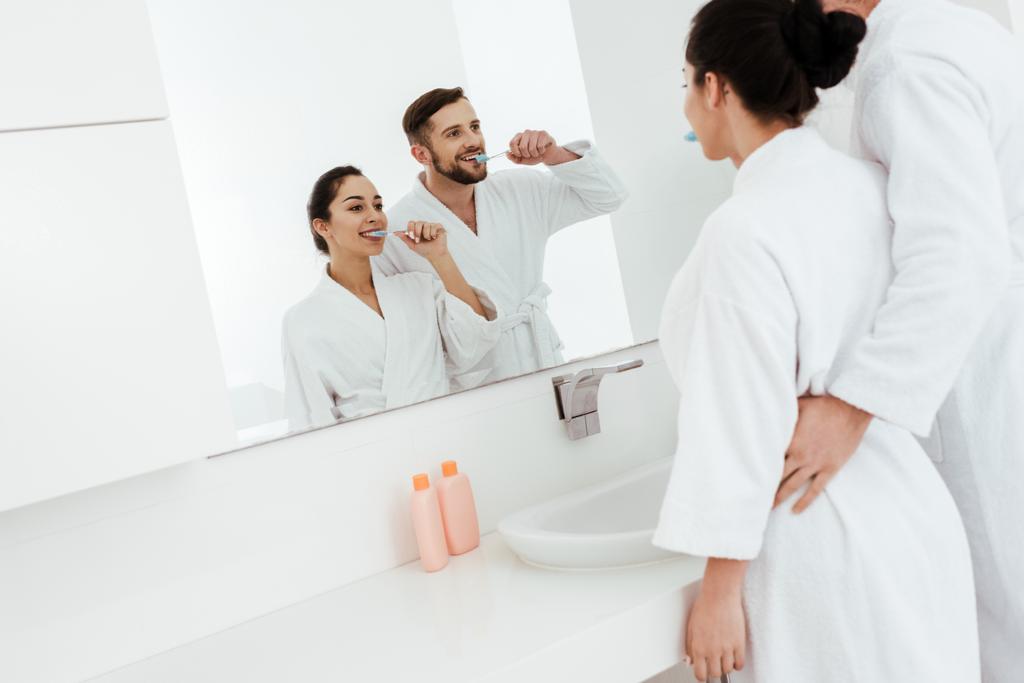陽気なカップルは、バスローブに立っている間、鏡を見て、歯を磨く  - 写真・画像