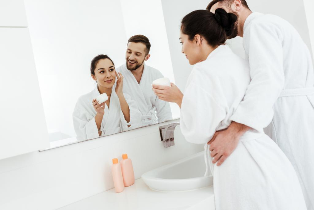 alegre hombre mirando hermosa novia aplicando crema facial en baño
  - Foto, imagen