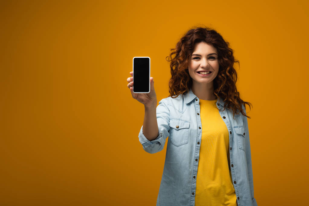 веселая рыжая женщина держит смартфон с пустым экраном на оранжевом
  - Фото, изображение